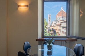 Un coin repas avec une table en verre, des chaises et une plante en pot. Une fenêtre offre une vue sur un bâtiment historique doté d’un dôme et d’un clocher.