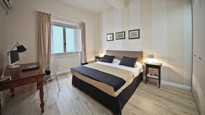 A hotel room with a large bed, a desk with a computer, a chair, a bedside table with a lamp, and a window with curtains. - Borgo dei Greci (Florence)