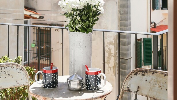 Un balcon avec une table, un vase rempli de fleurs et deux tasses dotées d’anses rouges.