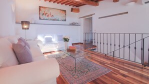 Un salon spacieux avec un canapé blanc, une table basse en verre et un tapis à motifs. Un tableau est accroché au mur, et un escalier avec une rampe en métal est visible au fond.