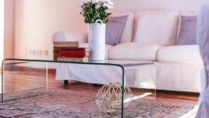 Un salon avec un canapé blanc, une table basse en verre, un vase blanc orné de fleurs et un tapis à motifs.
