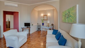 A living room with a white sofa, a coffee table, and a TV. There is a dining table with chairs and a lamp. An arched doorway leads to another room.