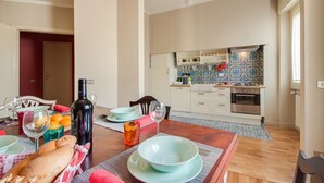 A dining area with a wooden table set for a meal, a kitchen in the background with white cabinets and a colorful backsplash, and a red door leading to another room.