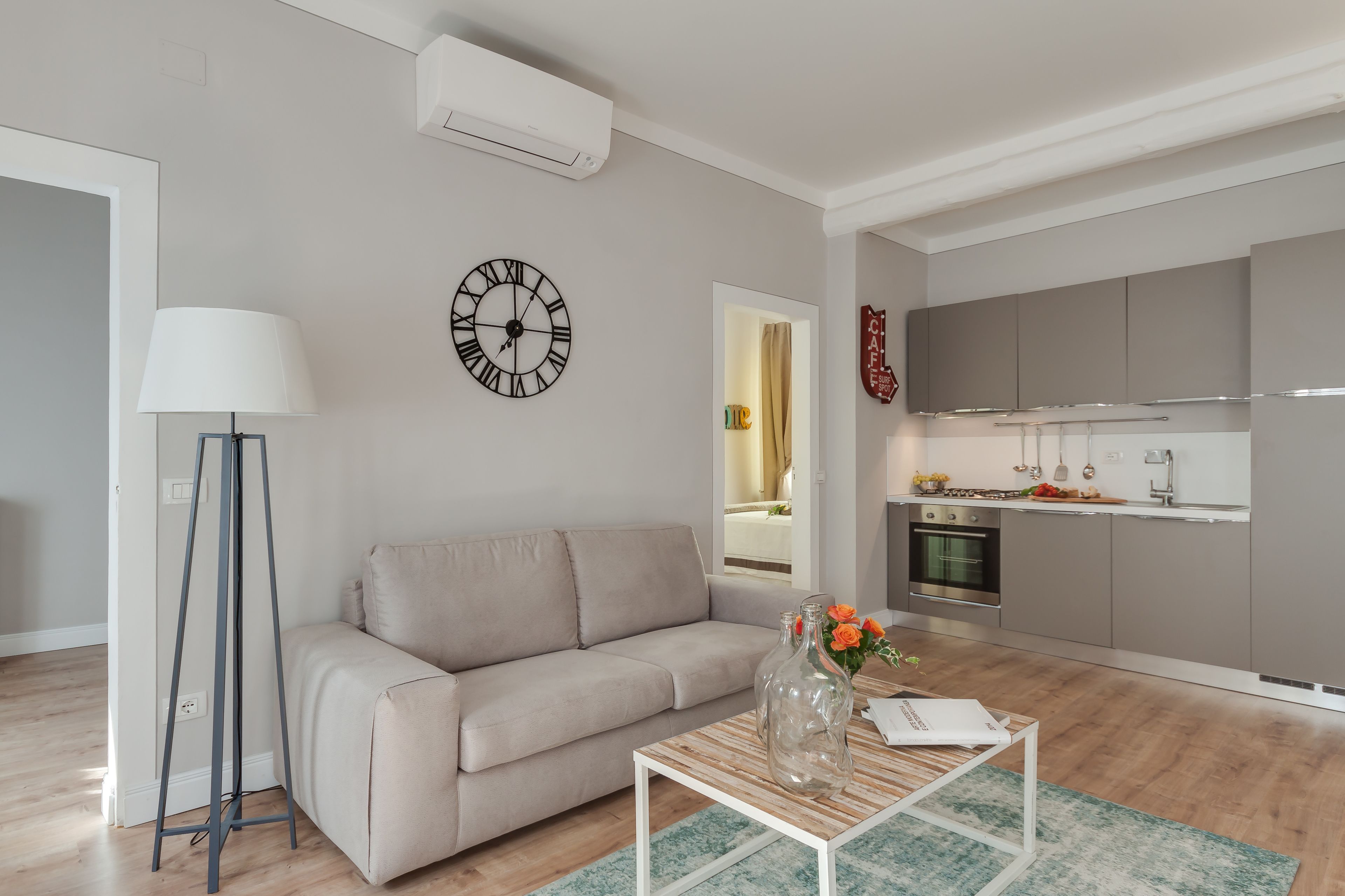 A modern living room with a sofa, a coffee table with a vase, a floor lamp, and a kitchen in the background.
