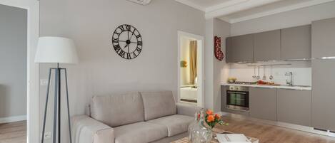 A modern living room with a sofa, a coffee table with a vase, a floor lamp, and a kitchen in the background.