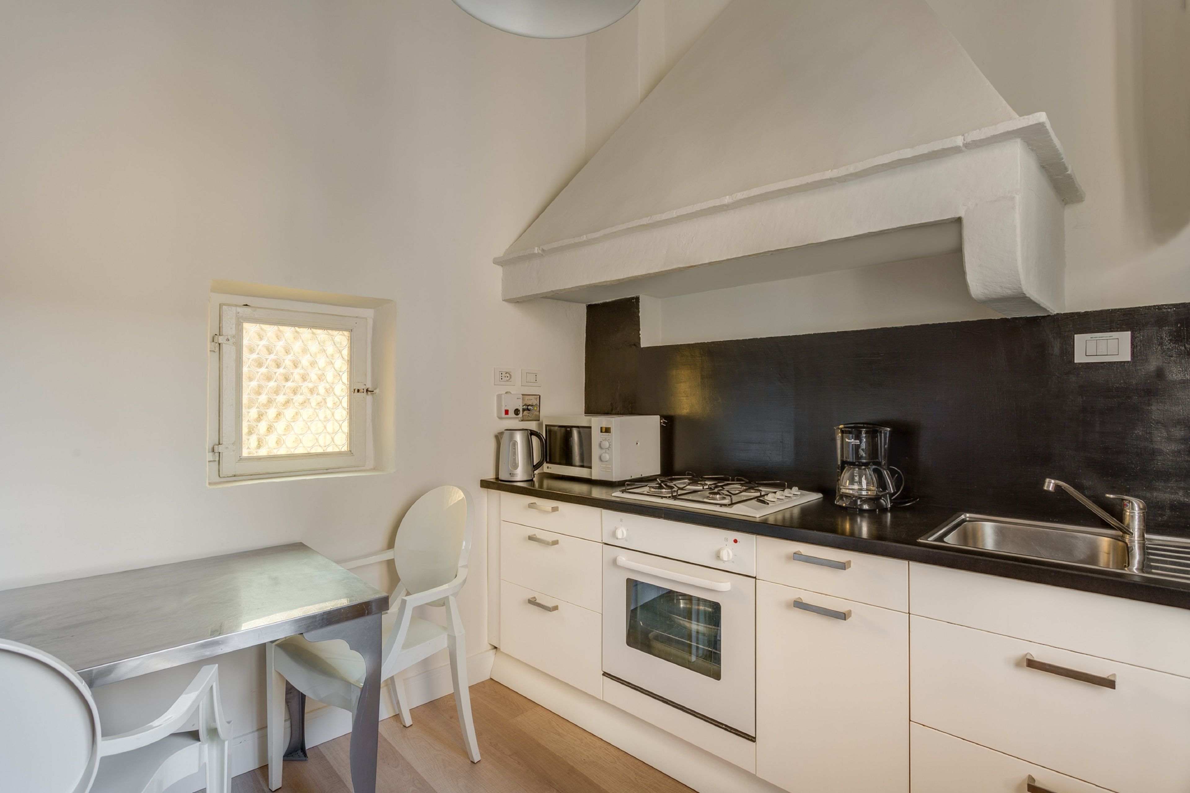 Een compacte keuken met een gasfornuis, magnetron, koffiezetapparaat en een kleine oven. Er staat een witte stoel bij een tafel met een lichtgekleurd aanrecht.