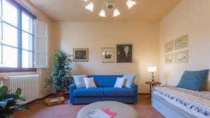 A living room with a blue sofa, a round ottoman, a small table with a lamp, and a bed with a patterned cover. There are framed pictures on the wall and a potted plant. - House in Florence Blue (Florence)