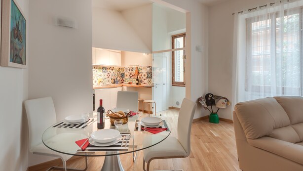 A modern living room with a glass dining table, white chairs, a beige sofa, and a kitchen in the background. - Liberta Loft (Florence)