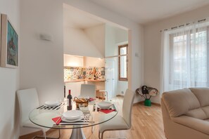 Un salon moderne avec une table à manger en verre, des chaises blanches, un canapé beige et une cuisine en arrière-plan.