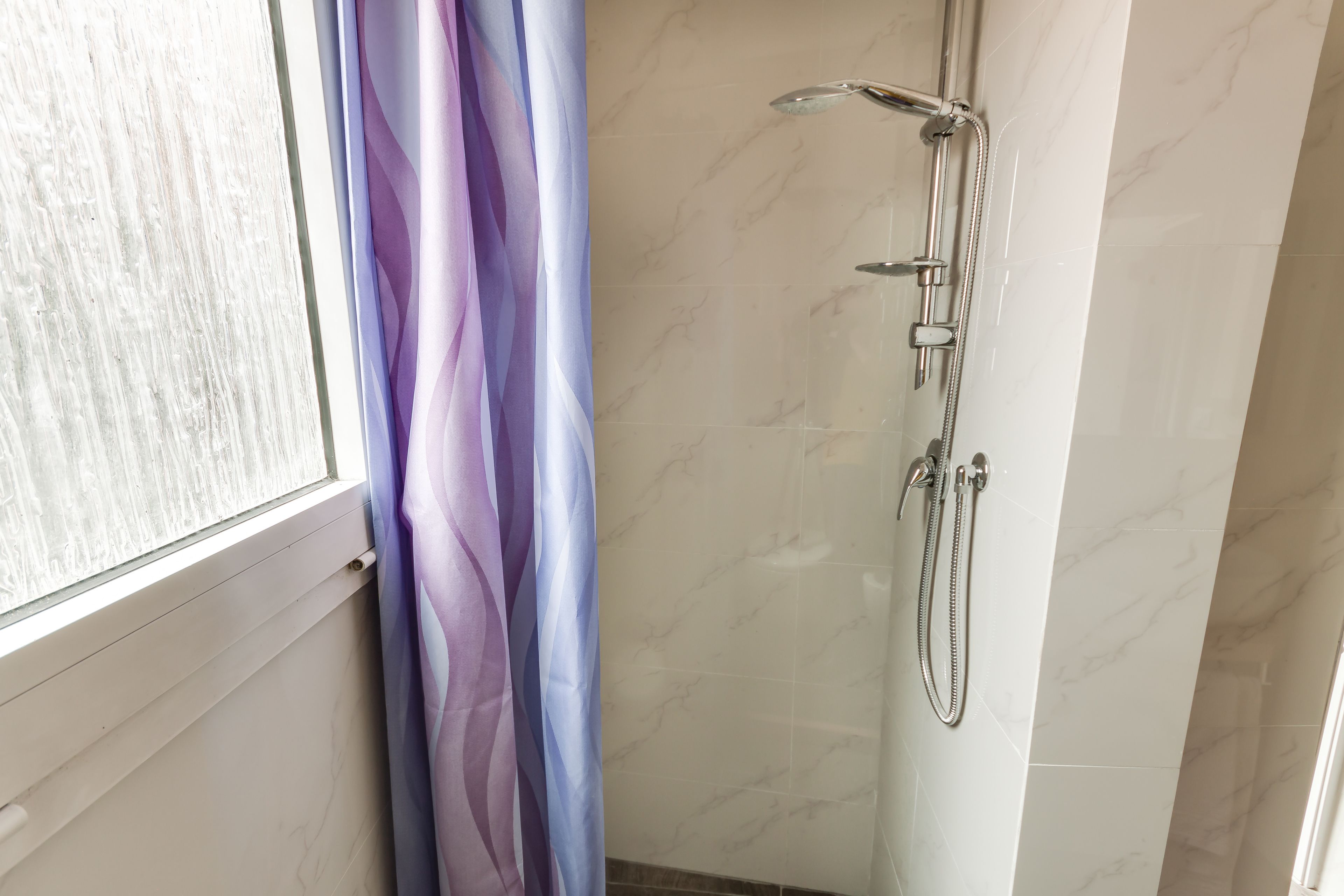 A bathroom with a shower, a window with a white frame, and a curtain with a purple and blue pattern.