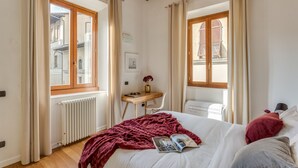 A bedroom with a bed, a desk, and a chair. There are windows with curtains, a radiator, and a wooden ceiling. - Luna Chic (Florence)