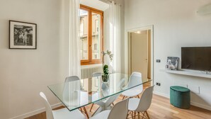 A modern dining area with a glass table and white chairs, a framed photo on the wall, and a view of a building through a window with white curtains. - Luna Chic (Florence)