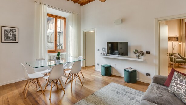 A spacious living area with a glass dining table, white chairs, a grey sofa, and a TV mounted on the wall. - Luna Chic (Florence)
