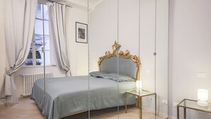A bedroom with a bed, a bedside table, a mirror, and a window with curtains. - Parione (Florence)