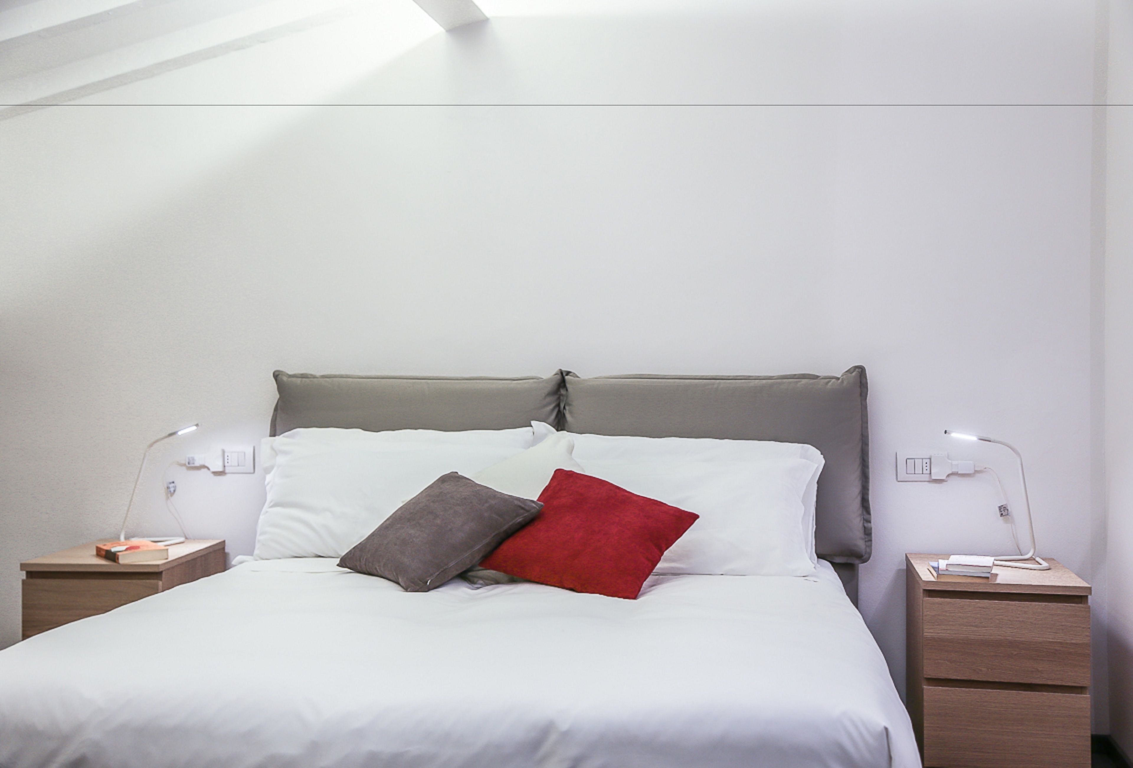 A neatly made bed with white bedding and three pillows in different colors. There are two bedside tables with lamps and a small drawer. The room has a modern, minimalist design with white walls and a slanted ceiling.