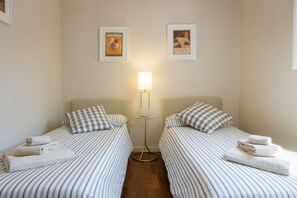 Una habitación de hotel con dos camas individuales, cada una con ropa de cama a rayas y almohadas cuadriculadas. Hay una lámpara de pie entre las camas y dos carteles enmarcados en la pared.