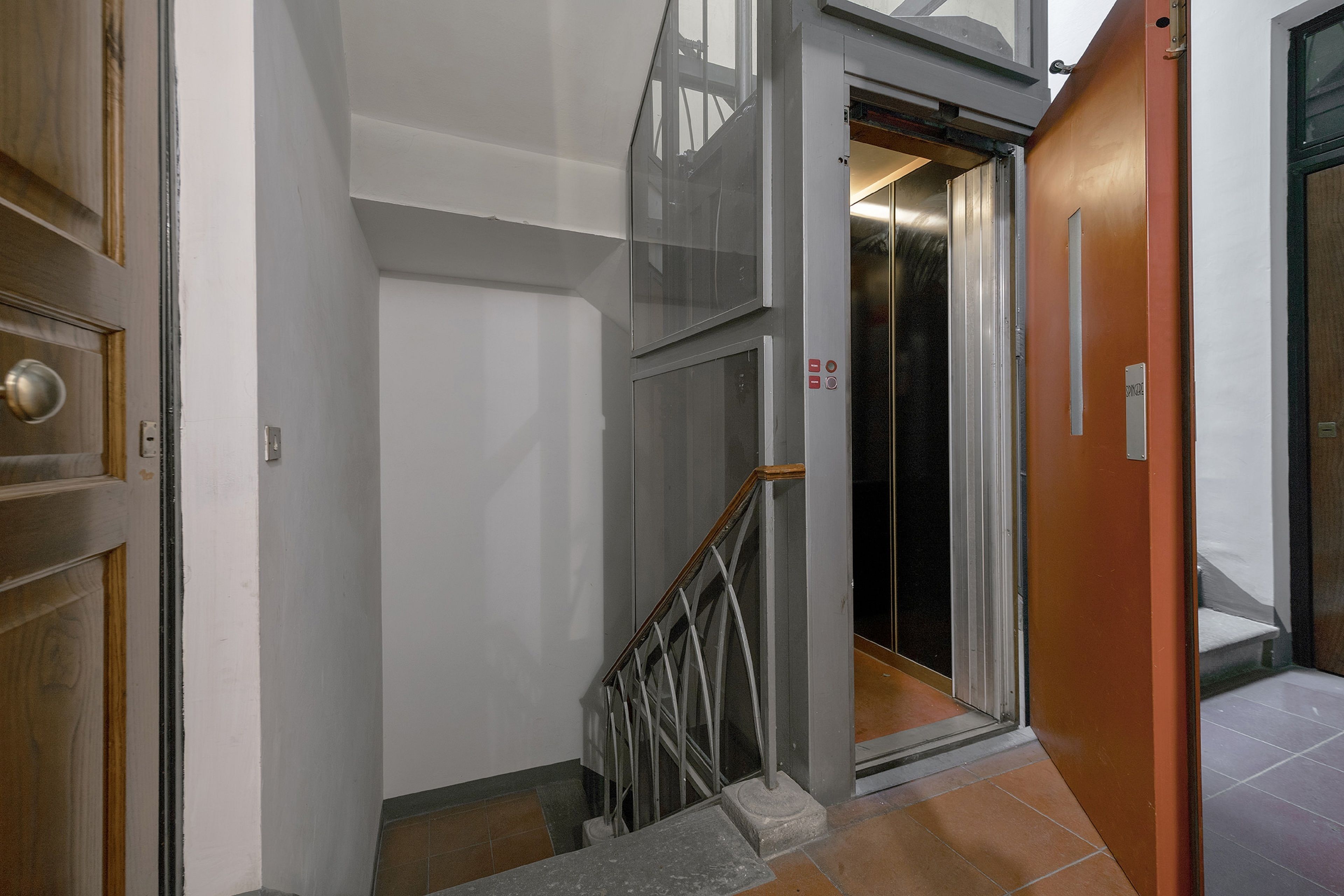 A modern elevator with a metal handrail and a glass door, leading to a room with a wooden door and a white wall.
