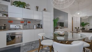 Een moderne keuken met witte kastjes, een glazen eettafel en witte stoelen. Er staan planten op de kastjes en er hangt een lamp.