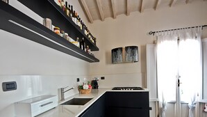 A modern kitchen with a white countertop, black cabinets, and a built-in stove. There are shelves above the counter holding various bottles and a basket with flowers. - Pergola in Palazzo Leopardi (Florence)