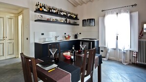 A dining area with a table set for four, a kitchen with wine bottles on shelves, and a window with curtains. - Pergola in Palazzo Leopardi (Florence)