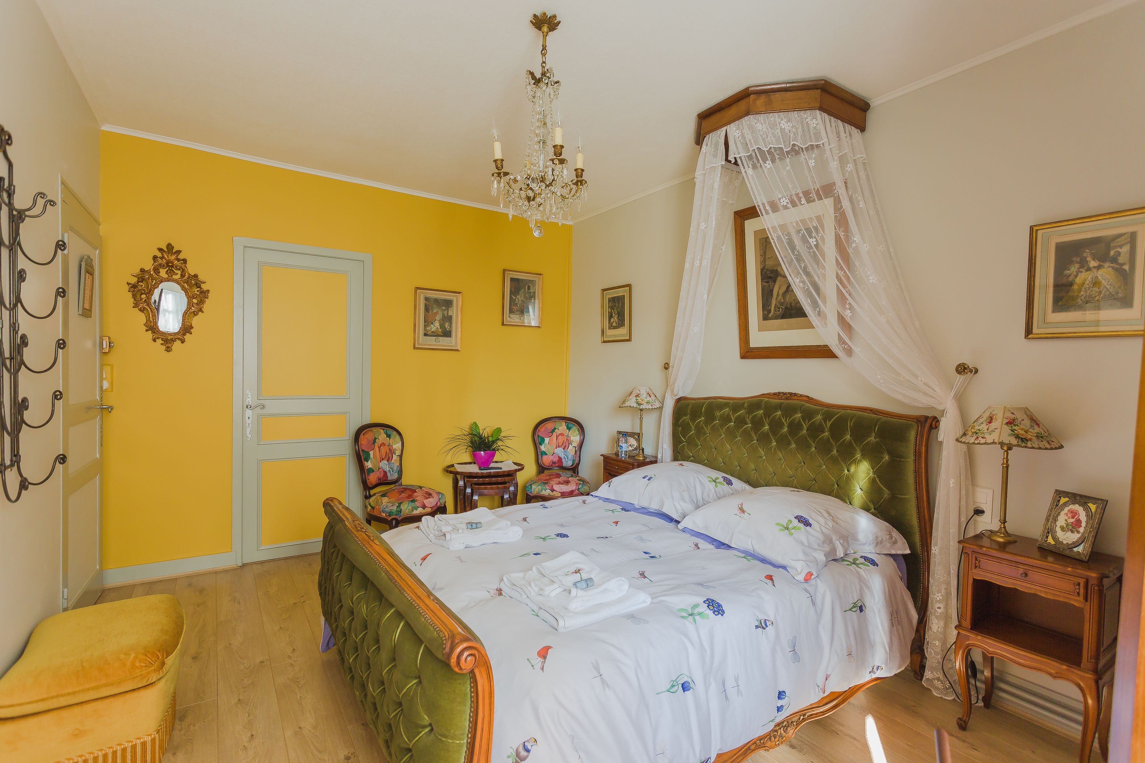 Een slaapkamer met een bed, een kroonluchter, een hemelbed, een houten bankje, een stoel, een kleine tafel en een fotolijst.