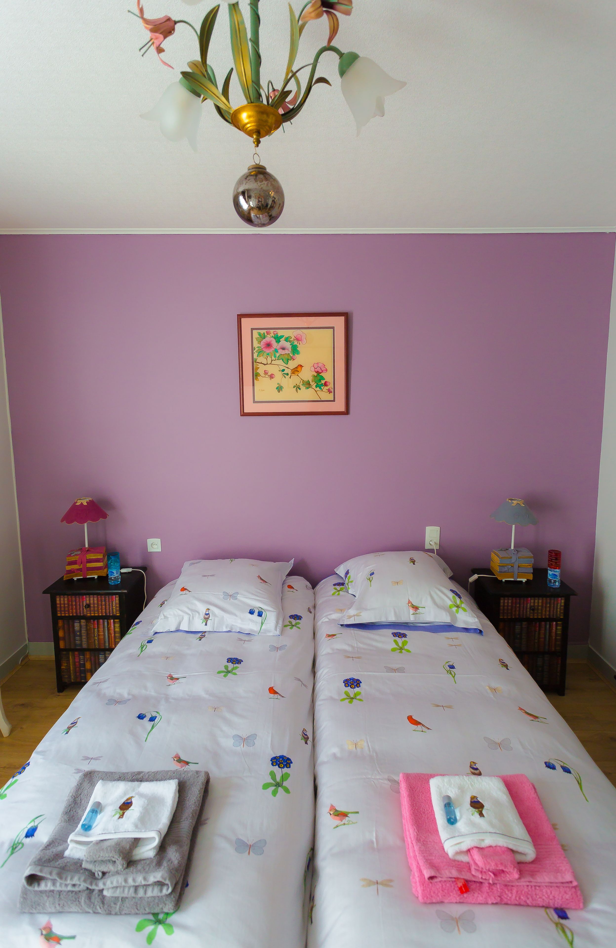 A bedroom with two beds, purple walls, a floral painting, and a chandelier.