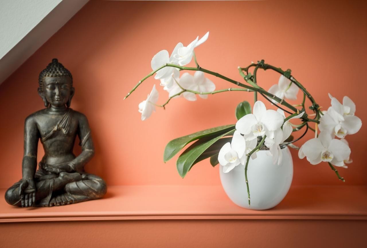 Een Boeddhabeeld en een vaas met witte orchideeën op een plank.