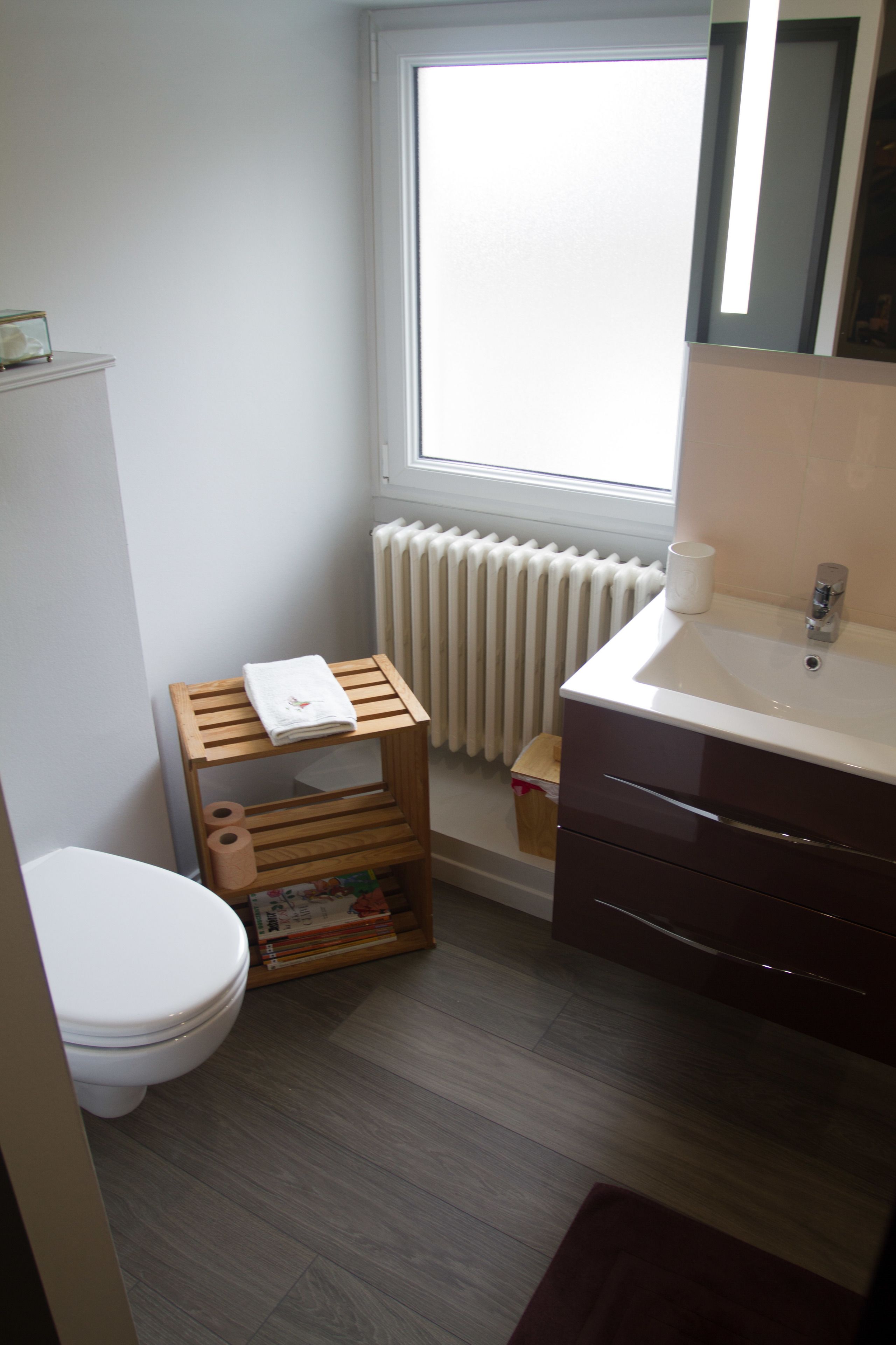A compact bathroom with a toilet, a wooden shelf with a towel, a small trash bin, and a window providing natural light.