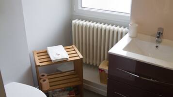 A compact bathroom with a toilet, a wooden shelf with a towel, a small trash bin, and a window providing natural light.