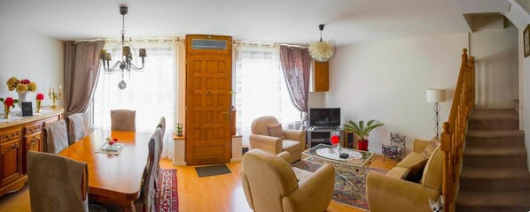 Una sala de estar con una mesa de comedor de madera, sofás color beige, un televisor y una escalera.