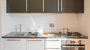 Eine moderne Küche mit dunklen Schränken, Edelstahlgeräten und einer Arbeitsplatte mit Spüle, Weingläsern und einem Schneidebrett mit Tomaten.