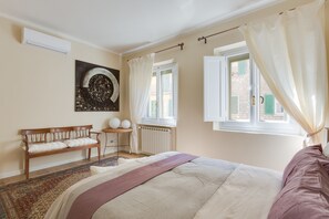 A bedroom with a bed, a bench, a small table, and a window with curtains. - Ponte Vecchio Balcony (Florence)
