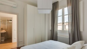 Un dormitorio con cama, ventana con cortinas, radiador y puerta que da al baño.