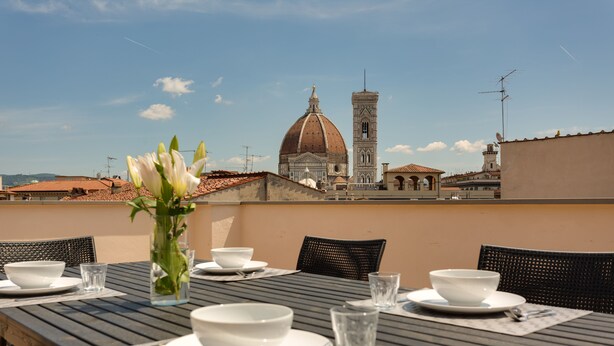Una mesa de comedor dispuesta con cuencos, platos y vasos blancos, con un jarrón de flores en el centro. Al fondo, se observa una vista de un edificio histórico con una cúpula y un campanario.
