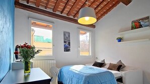 Un dormitorio con cama, escritorio y silla. Hay ventanas, un ventilador de techo y una estantería con una planta.