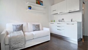 Una sala de estar moderna con un sofá blanco, un plaid decorativo y una pequeña planta en maceta sobre un estante blanco.