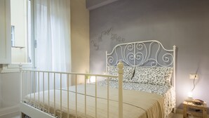A bedroom with a white metal bed frame, a patterned bedspread, a wall with a decorative design, a bedside table with a lamp, and a window with curtains. - San Lorenzo Chic (Florence)