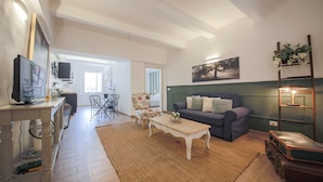 A living room with a sofa, coffee table, and a television. There is a dining area with chairs and a table. The room has a large window and a painting on the wall. - San Marco Suite III (Florence)