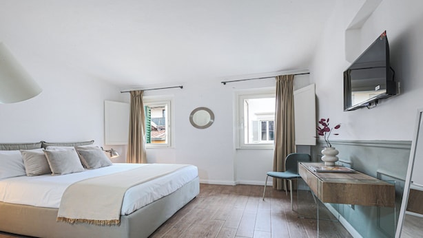 A bedroom with a bed, a desk, a chair, a mirror, and a window with curtains. - San Marco Suite VII (Florence)