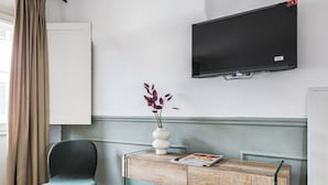 A room with a TV mounted on the wall, a wooden desk with a vase and magazine, a chair, and a window with curtains. - San Marco Suite VII (Florence)