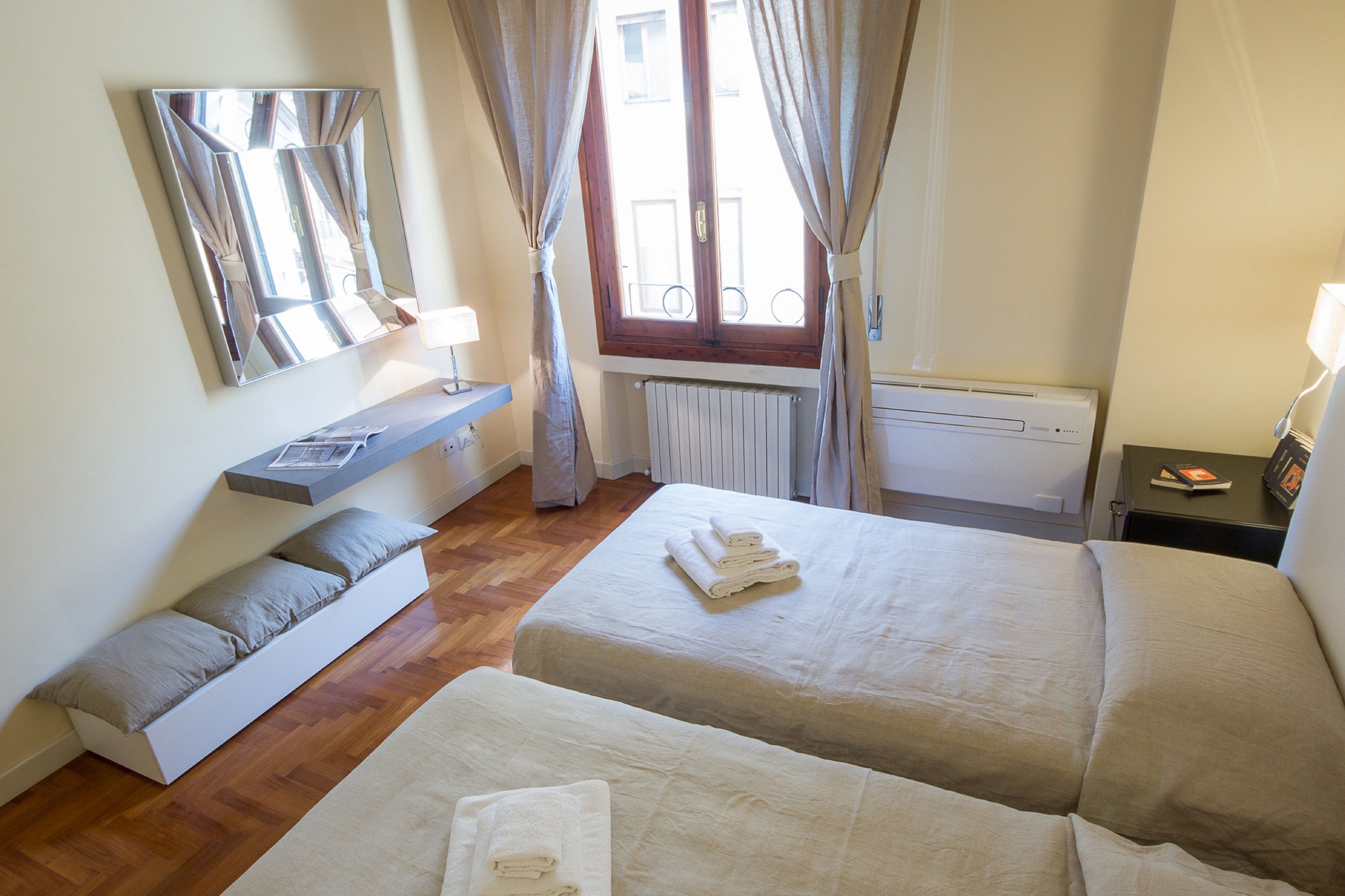 A hotel room with two beds, a mirror, a window with curtains, a radiator, and a bedside table with a lamp.
