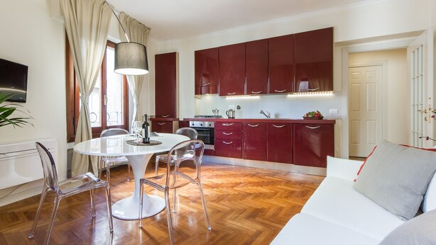 A modern kitchen with red cabinets, a white dining table with clear chairs, and a large window with curtains. - Santa Maria View (Florence)
