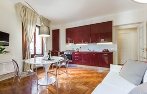 A modern kitchen with red cabinets, a white dining table with clear chairs, and a large window with curtains.