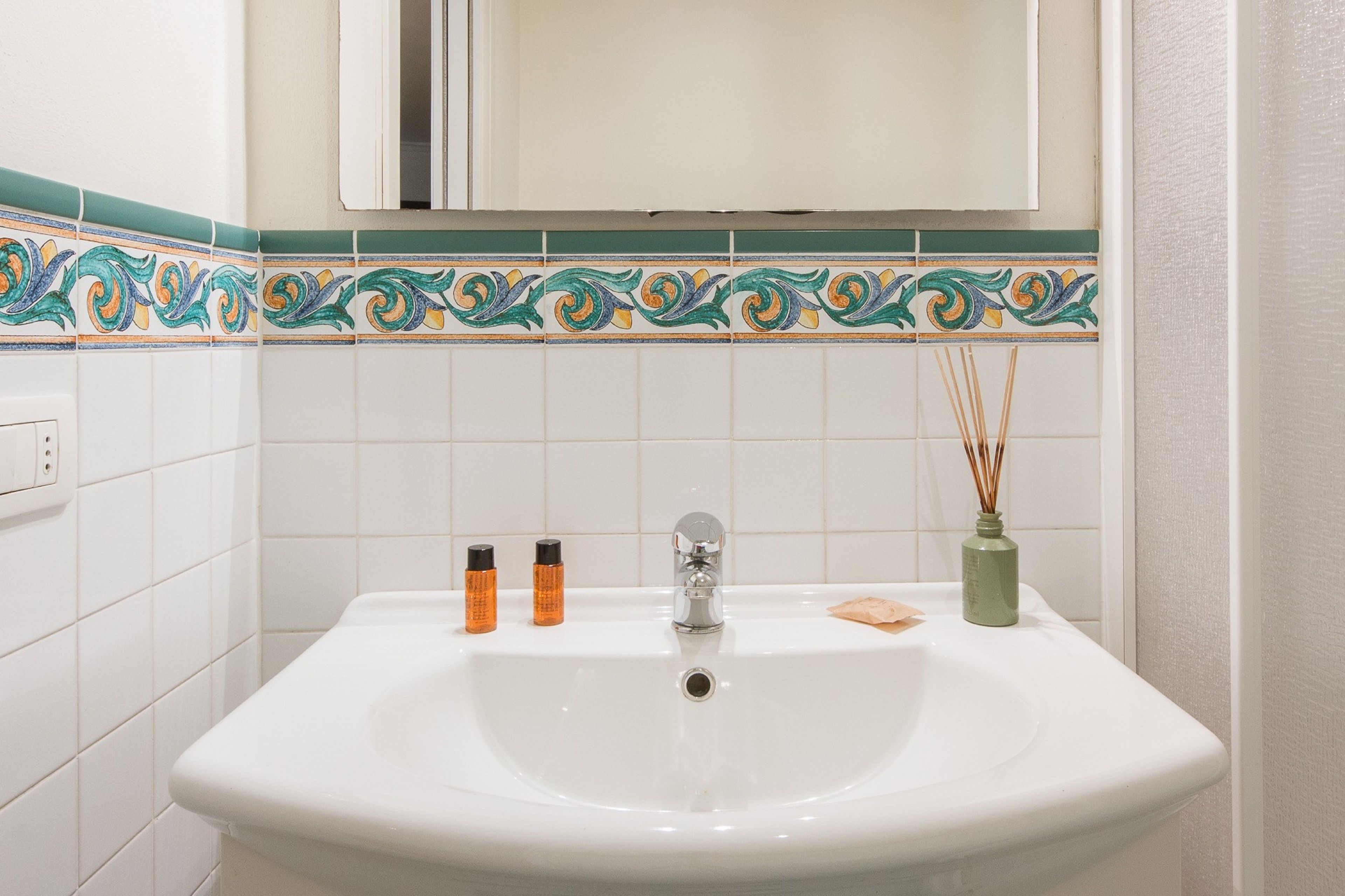A bathroom with a white sink, two bottles of toiletries, a green vase with reed sticks, and a decorative tile border.