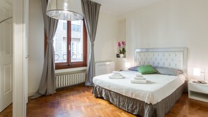 A bedroom with a large bed, a window with curtains, a radiator, and a bedside table with a lamp. - Santa Maria View (Florence)