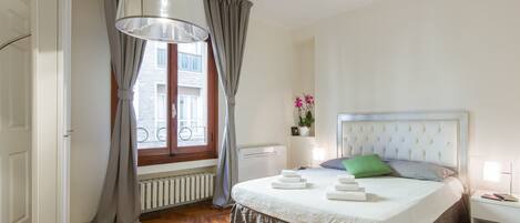 Un dormitorio con una cama grande, una ventana con cortinas, un radiador y una mesita de noche con una lámpara.