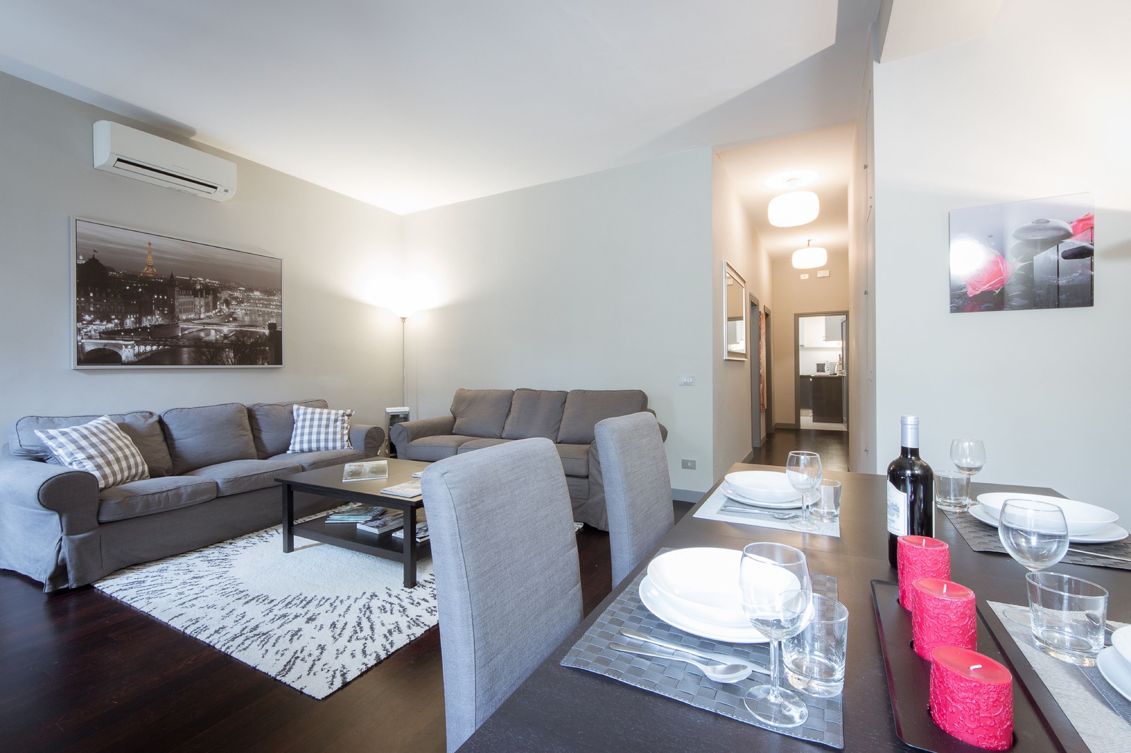 A modern living room with a gray sofa, a coffee table, and a dining area set with plates, glasses, and a wine bottle.