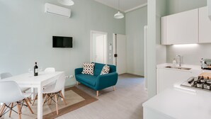 Een moderne keuken met witte kasten en een ingebouwde kookplaat, een eethoek met een witte tafel en stoelen, en een blauwgroene bank met gekleurde kussens.