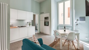 Een moderne keuken met witte kasten, een eethoek met een witte tafel en stoelen, en een teal-kleuren bank.