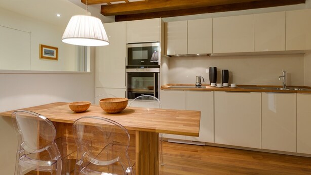 Una cocina moderna con vigas de madera, armarios blancos y una mesa de comedor de madera con sillas transparentes.